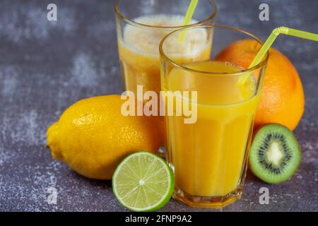Jus d'orange fraîchement pressé et smoothies à la banane et à l'orange dans de grands verres avec une paille entourée de fruits sur un fond de béton gris. Le con Banque D'Images
