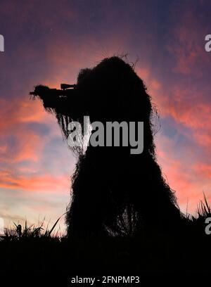 Paintball Sniper en Full Ghillie costume camouflage silhoueté contre un ciel de coucher de soleil sur une colline avec son arme tirée, visant et prêt à tirer Banque D'Images