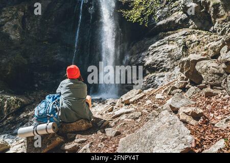 Femme avec un sac à dos en chapeau rouge vêtu de vêtements de randonnée actifs assis près de la cascade de la rivière de montagne et appréciant la puissance de nature éclabousse. Banque D'Images