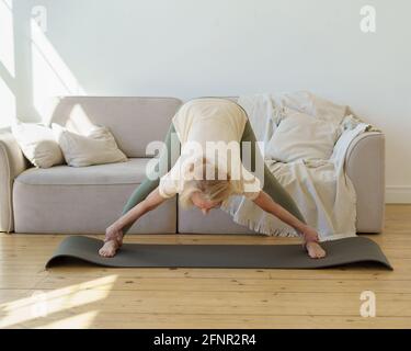 Active femme sénior des années 60 dans les sports vêtements pratiquant le yoga de chien face vers le bas pose tout en faisant de l'exercice à la maison. Mature flexible dame faisant large leged en avant courbe ou Prasarita Padottanasana posture Banque D'Images