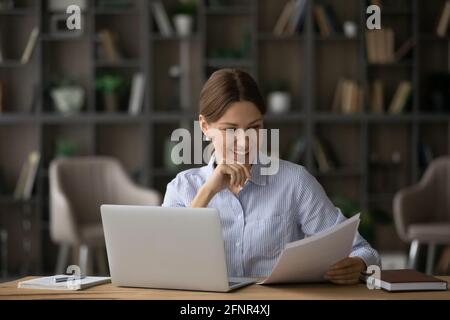 Une femme millénaire satisfaite lit les documents papier avant l'écran d'ordinateur Banque D'Images