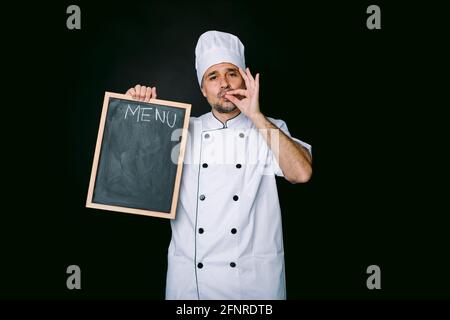 Chef cuisinier portant une veste de cuisine et un chapeau, tient une piazarra avec le menu et fait un bon geste de nourriture, sur fond noir Banque D'Images