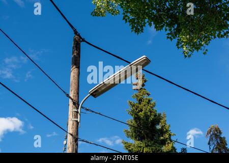 Un lampadaire de rue avec des fils à côté d'arbres verts sur une rue. Poteau en bois avec une lanterne et des câbles électriques contre un ciel bleu. Banque D'Images