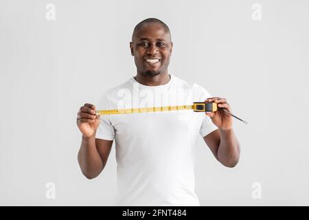 Joyeux homme de main afro-américain ou constructeur dans la tenue de t-shirt blanc mètre-ruban et sourire à la caméra Banque D'Images