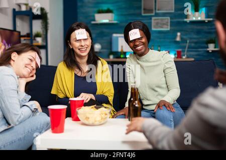 Des amis de course mixtes jouant deviner qui jeu avec des papiers collants fixés sur le front. Groupe de personnes multiraciales qui s'amusent, riant ensemble tout en étant assis sur un canapé dans le salon tard dans la nuit. Banque D'Images