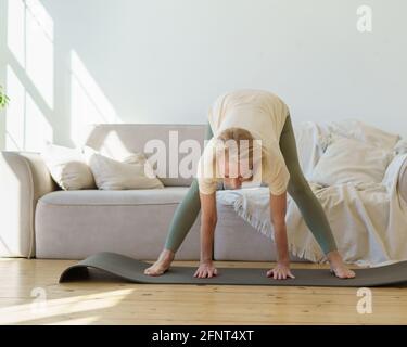 Active femme sénior des années 60 dans les sports vêtements pratiquant le yoga de chien face vers le bas pose tout en faisant de l'exercice à la maison. Mature flexible dame faisant large leged en avant courbe ou Prasarita Padottanasana posture Banque D'Images