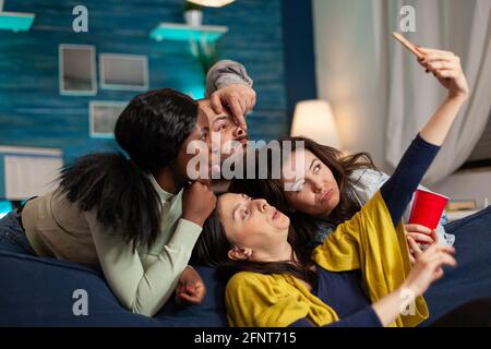 Groupe de personnes de race mixte prenant des photos avec le téléphone tout en étant assis sur un canapé dans le salon passant du temps ensemble. Amis multiraciaux poster selfie sur le partage d'Internet avec d'autres personnes. Banque D'Images