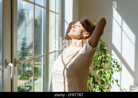 Nouvelle journée. Belle heureuse femme mûre s'étirant le corps et souriant tout en se tenant avec les yeux fermés dans les rayons du soleil qui brillent à travers la fenêtre le matin ensoleillé à la maison. Concept d'optimisme et de bonheur Banque D'Images