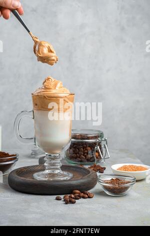 Une boisson avec de la mousse de café, du lait et de la glace et une cuillère avec de la mousse dans la main d'une femme sur fond gris avec un espace à copier. Café Dalgona. Banque D'Images