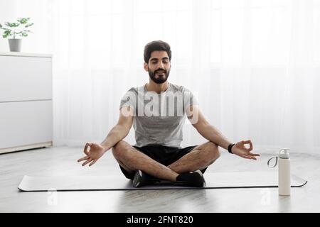 Homme millénaire à barbe arabe gai assis sur un tapis en position lotus, sur le sol avec une bouteille d'eau Banque D'Images