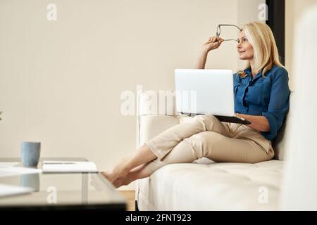 Femme d'affaires pensive et mûre souriant loin de chez elle, assise sur un canapé dans un appartement moderne Banque D'Images
