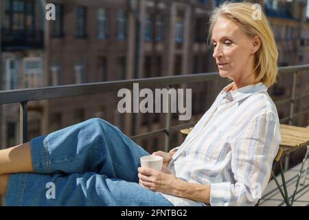 Le matin. Belle femme adulte rêveuse des années 60 dans une tenue décontractée se détendre sur le balcon à la maison, commencer la journée avec le café. Bonne femme âgée assise sur la terrasse et regardant la rue ensoleillée Banque D'Images