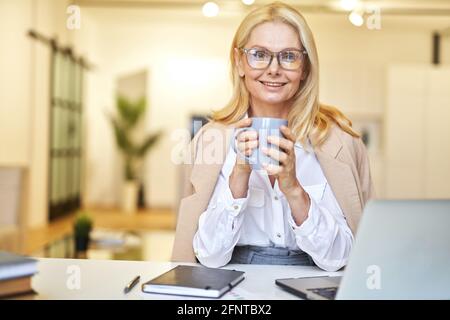 Belle femme d'affaires mûre dans des verres souriant à l'appareil photo et à boire café tout en travaillant sur l'ordinateur portable dans le bureau Banque D'Images