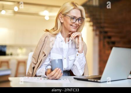 Femme d'affaires souriante et mûre dans des verres utilisant un ordinateur portable et buvant du café tout en faisant quelques documents au bureau Banque D'Images