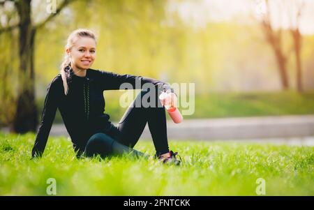 Jeune fille blanche assez soif blonde de race blanche en costume se reposant après course sportive dans le stade de football assis sur l'herbe buvant l'eau et le sourire enjoies Banque D'Images