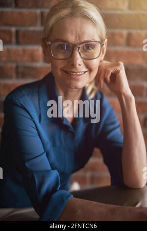 Portrait d'une femme d'affaires blanche, mûre et confiante, en lunettes souriant à la caméra tout en posant, assise devant un mur de briques Banque D'Images