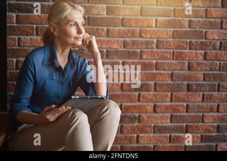 Femme blanche mûre paisible et blonde qui a l'air attentionnés, tenant un Tablet pc tout en étant assis devant un mur de briques à la maison Banque D'Images