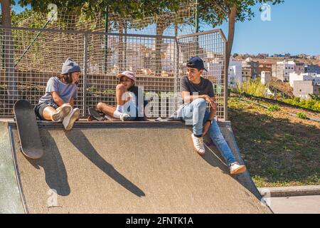 Trois jeunes patineurs caucasiens assis sur un obstacle dans le parc de skate, avec leurs patins de côté, traîner ou se reposer, parler et rire très joie Banque D'Images