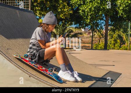 Une fille de race blanche assise sur une rampe de skateboard avec sa planche à côté d'elle, tout en regardant son téléphone mobile Banque D'Images
