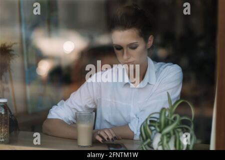 Belle jeune fille s'assoit dans un café près de la fenêtre derrière un verre avec une tasse de café et envoie un message sur le smartphone. Photo à travers verre avec Banque D'Images