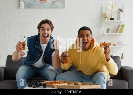 KIEV, UKRAINE - 22 MARS 2021 : amis interraciaux assis sur un canapé avec des bouteilles de bière et de pizza Banque D'Images