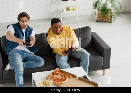 KIEV, UKRAINE - 22 MARS 2021: Vue en grand angle des amis interraciaux jouant émotionnellement le jeu vidéo avec des joysticks et appréciant la pizza sur le canapé en Mo Banque D'Images