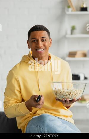 homme afro-américain souriant en cliquant sur les chaînes avec la télécommande de télévision et manger du pop-corn dans un loft moderne Banque D'Images