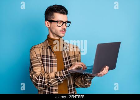 Photo de jeune homme sérieux et élégant programmeur en lunettes travailler sur un ordinateur portable isolé sur un arrière-plan bleu Banque D'Images