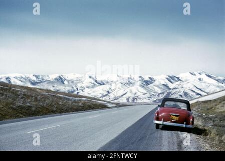 Longue route droite avec 1952 voitures Red Plymouth convertibles garées au bord de la route avec une vue sur la chaîne de montagnes Rocheuses enneigées, États-Unis dans les années 1950 Banque D'Images