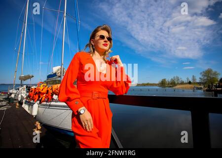 Belle femme dans le costume rouge vif sur le fond du yacht. La blonde en lunettes de soleil sur le fond du yacht et le ciel bleu. Banque D'Images