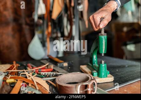 Main d'homme méconnaissable utilisant un perforateur travaillant avec du cuir dans un atelier avec d'autres outils. Banque D'Images