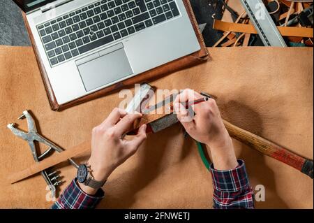 Point de vue artisan méconnaissable prenant des mesures d'une sangle en cuir tout en travaillant dans son atelier. Ordinateur portable et de nombreux outils à bord. Banque D'Images