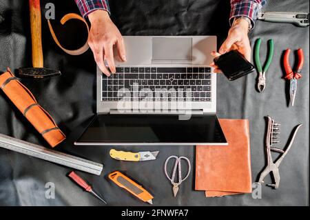 Vue de dessus Homme méconnaissable tenant un smartphone et utilisant un ordinateur portable travaillant dans un atelier avec un bureau rempli d'outils de travail pour les articles en cuir. Banque D'Images