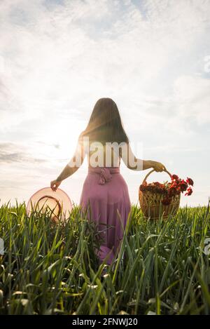 Belle fille du dos au coucher du soleil, dans un pays de blé vert, tenant un chapeau dans sa main gauche et un panier avec des fleurs dans sa main droite Banque D'Images