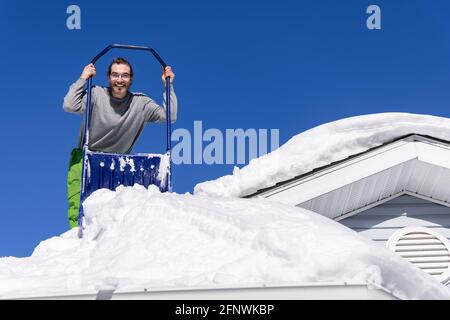 Vue en grand angle depuis le dessous d'un jeune homme posant avec une pelle manuelle bleue depuis le toit d'une maison où il enlève des tas de neige fraîche. Banque D'Images