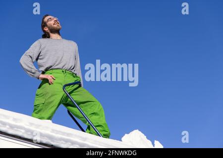 Portrait amusant de dessous d'un jeune homme debout sur le toit d'une maison, bras sur sa hanche et pelle bleue sur son côté, pendant une opération d'enlèvement de neige Banque D'Images