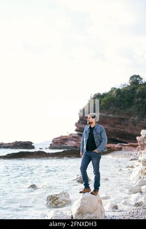 Homme en Jean et une veste en denim se tient à regarder la mer sur une grande pierre sur une plage de galets sur fond de montagnes rocheuses verdoyantes Banque D'Images