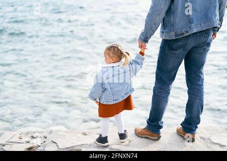 Papa dans une veste en denim et jeans tient une petite fille à la main tout en se tenant sur les pierres près de l'eau. Vue arrière Banque D'Images