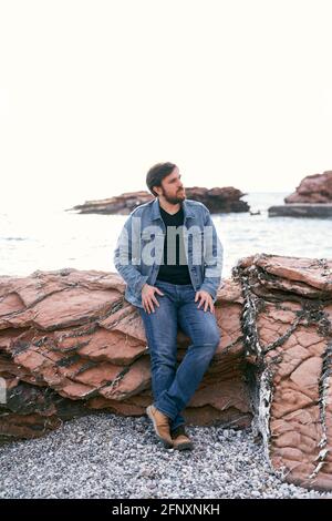 Homme pensif en Jean et une veste en denim se penche contre les pierres près de la mer et regarde loin Banque D'Images