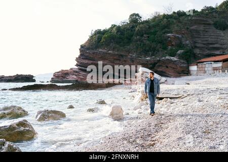 Un homme pensif en Jean et une veste en denim se promène une plage de galets regardant l'eau sur un fond de roches vertes Banque D'Images