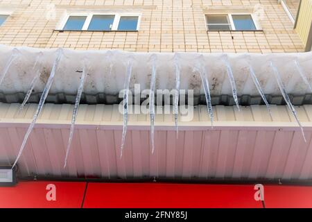De longues glaces pendent dangereusement du toit. Le mur du bâtiment est fini avec des carreaux de plastique rouge et blanc. Concept de conditions météorologiques anormales. Le nouvel an et Noël sont terminé. Banque D'Images