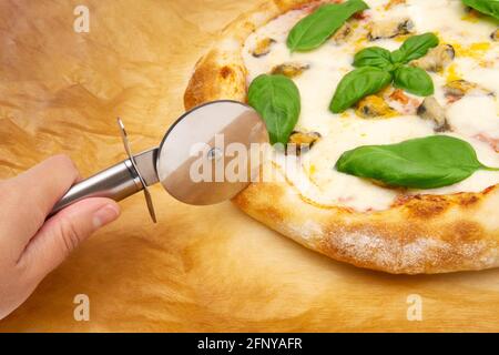 Main féminine commençant à couper une pizza napolitaine avec un coupe-pizza sur un papier parchemin Banque D'Images