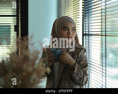 Portrait d'une jeune femme d'affaires musulmane qui se détend avec un café tout en regardant hors de la fenêtre Banque D'Images