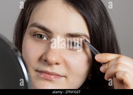 Une femme brune a plé ses sourcils devant un miroir. Correction de la forme du sourcil à l'aide d'une pince à épiler seule Banque D'Images