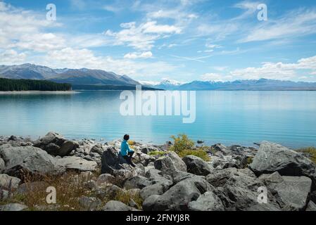 Femme assise sur les rives du lac Pukaki, bénéficiant de la vue sur le Mont Cook et les Alpes du Sud, sur l'île du Sud. Banque D'Images