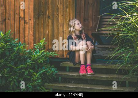 adorable fille blonde assise dans un escalier près d'un chalet en bois jardin verdoyant Banque D'Images