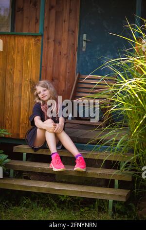 adorable fille blonde assise dans un escalier près d'un chalet en bois jardin verdoyant Banque D'Images