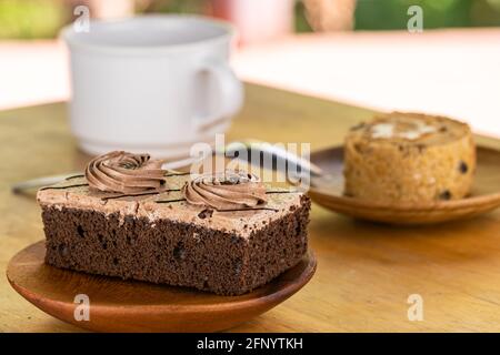 Mini-gâteau de crème au chocolat dans une assiette et une pièce en bois de pruner le gâteau éponge dans un plat en bois avec une tasse de café et cuillère en métal sur table en bois Banque D'Images