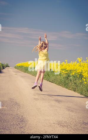 petite fille sautant parmi les champs de colza jaune en milieu rural vide chemin Banque D'Images