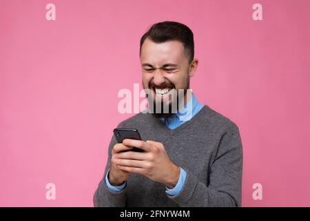 Gros plan sur un sourire joyeux heureux cool bien beau brunet unshaven jeune homme avec barbe portant un chandail gris et une chemise bleue isolée sur le rose Banque D'Images